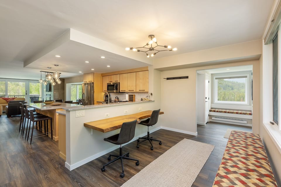 A modern, open-plan kitchen and dining area with wooden cabinets, a breakfast bar, dining table, and large windows allowing natural light to fill the space. A hallway leads to another room.