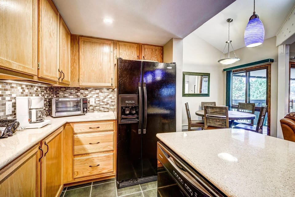 A kitchen with wooden cabinets, a black refrigerator, a microwave, and a dishwasher. A dining area with a table and chairs is visible in the background, with blue pendant lights above the counter.