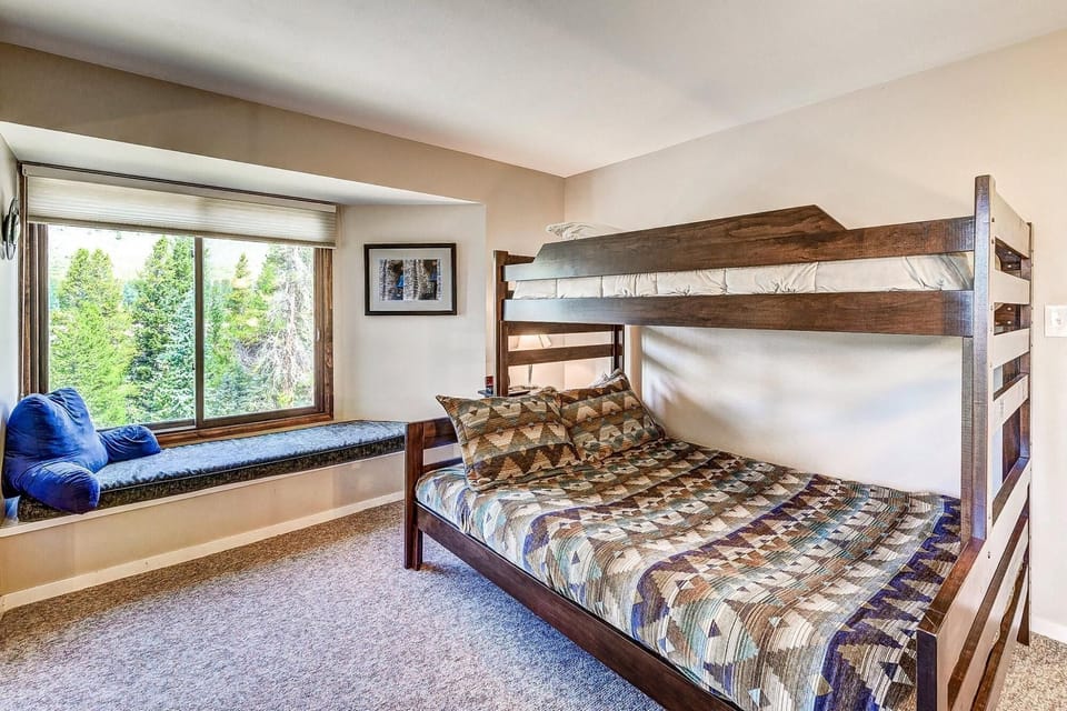 A bedroom featuring a dark wooden bunk bed with geometric-patterned bedding, a window seat with blue cushions, and a large window overlooking greenery.
