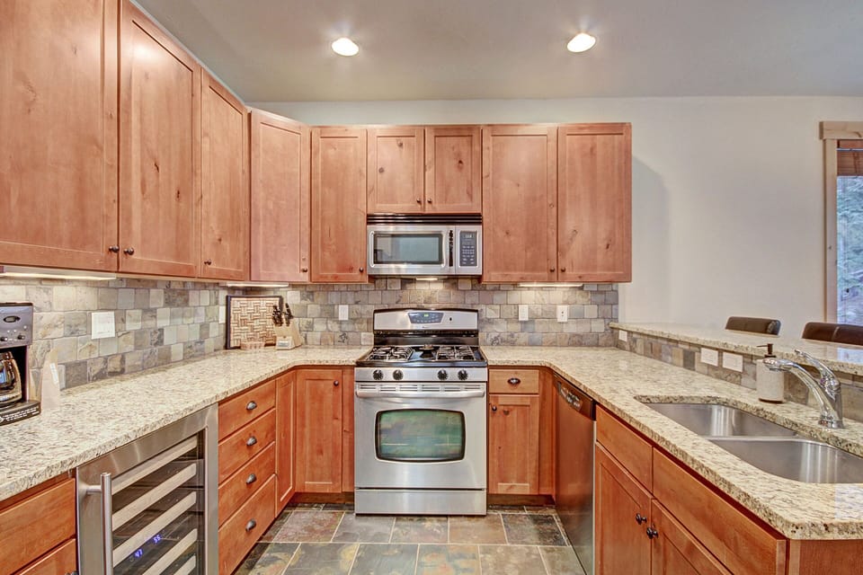 A modern kitchen with wooden cabinets, granite countertops, stainless steel stove, microwave, and dishwasher, a small wine fridge, coffee maker, and a double sink. The backsplash is tiled.