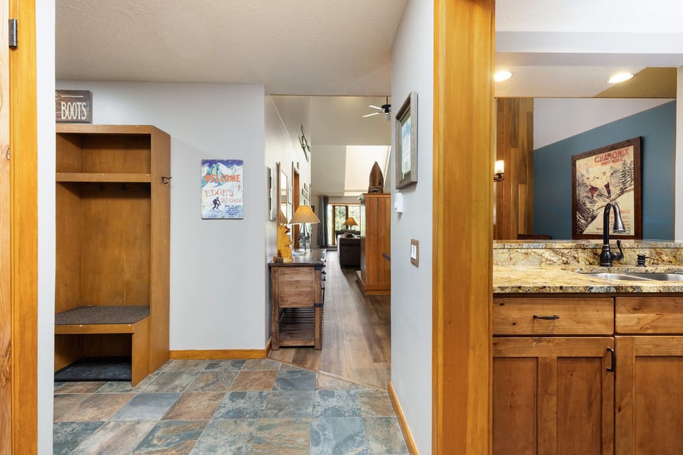 Interior view of a house featuring a small tiled mudroom on the left, leading to a hallway and a living area. To the right, there is a kitchen countertop with a sink and overhead cabinets.