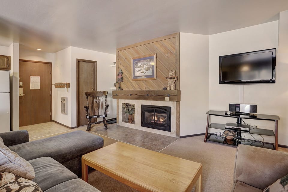 A cozy living room with a fireplace, a mounted TV, a wooden rocking chair, and a gray sectional sofa. A wooden coffee table sits in front of the sofa. The kitchen area is visible in the background.