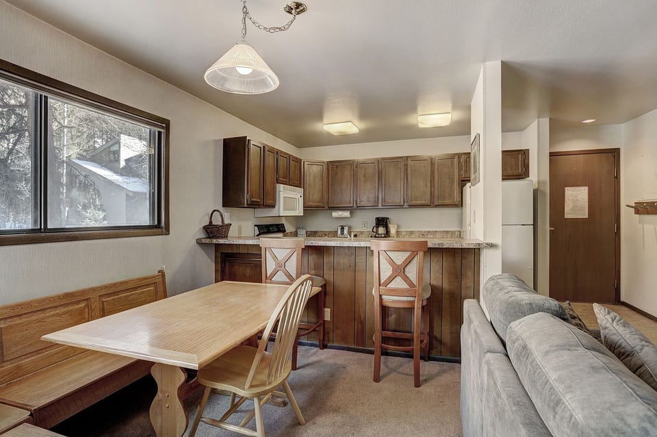 A small kitchen with light brown cabinets, a white refrigerator, a microwave, and a coffee maker. Adjacent is a light wood dining table with benches and chairs, next to a gray couch.