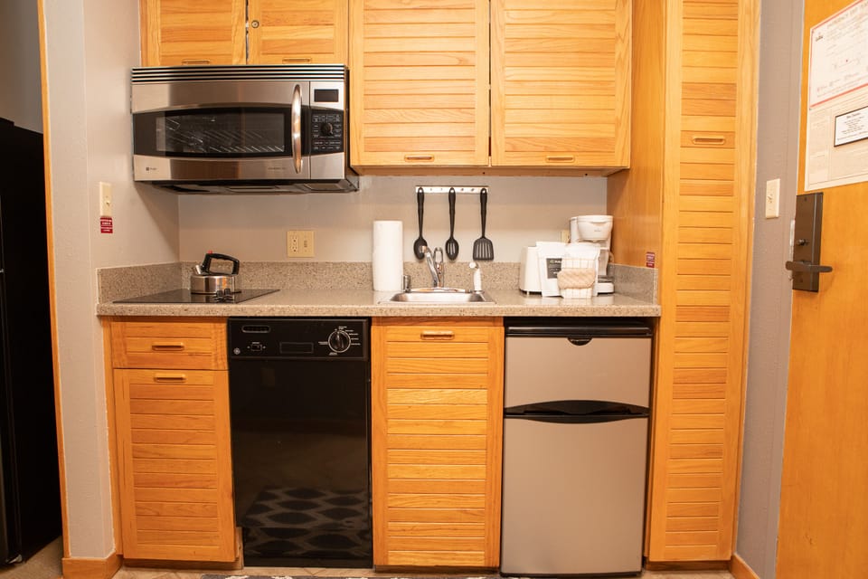 A small kitchen with wood cabinets, a microwave, electric stovetop, dishwasher, compact refrigerator, sink, and utensil holder on a granite countertop.