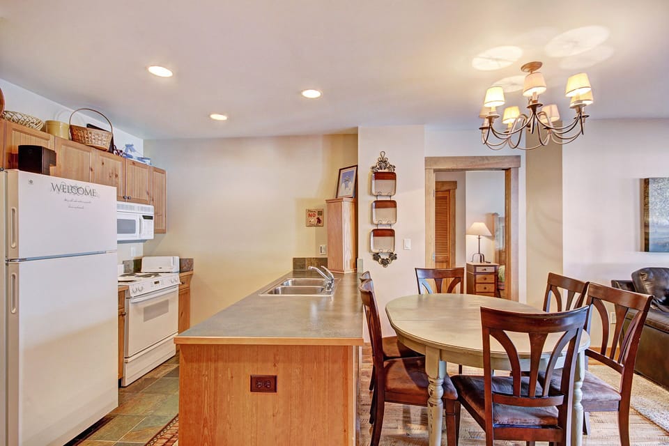 A kitchen with wooden cabinets, white appliances, an island with a sink, and a round dining table with six chairs under a chandelier. The room has tile flooring and a visible doorway to the next room.