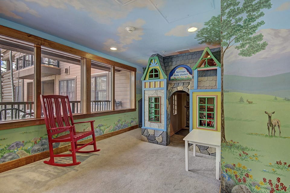 A children's playroom with a red rocking chair, a small playhouse, and walls painted with outdoor and nature scenes, including trees and a deer. Large windows fill one wall, providing natural light.
