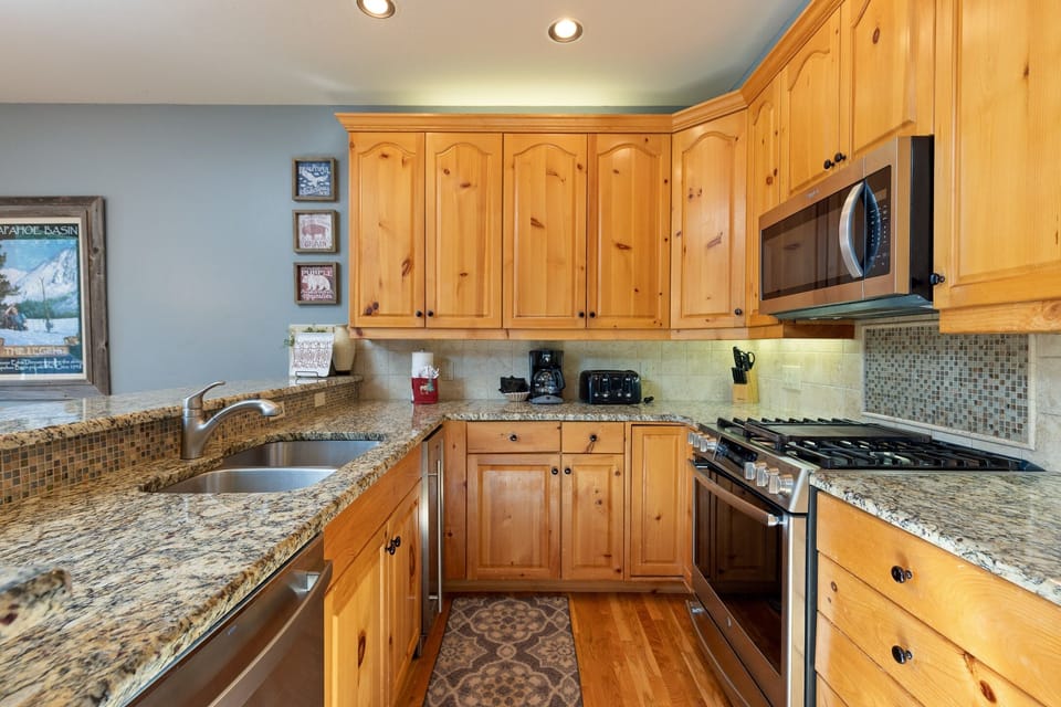 A kitchen featuring wooden cabinets, granite countertops, stainless steel appliances, a double sink, and a mosaic tile backsplash. There is a microwave above the stove and various kitchen items on the counter.