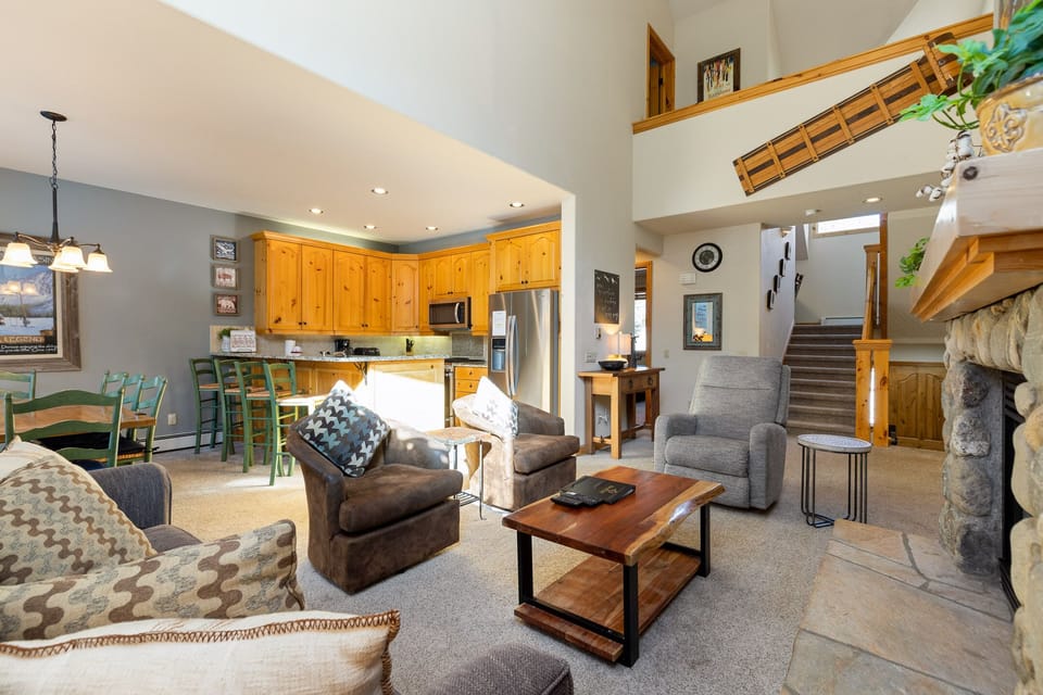 A spacious living area with a high ceiling features a cozy seating arrangement, a fireplace, a wooden coffee table, a kitchen with wooden cabinets, and an upstairs loft area.
