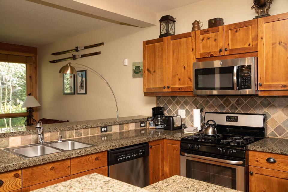A modern kitchen featuring wooden cabinets, granite countertops, a stainless steel oven, a microwave, and a dishwasher. A living area with a window view is partially visible in the background.