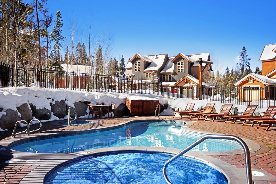 A view of an outdoor swimming pool and hot tub surrounded by snow with lounge chairs on the pool deck and houses in the background under a clear blue sky.