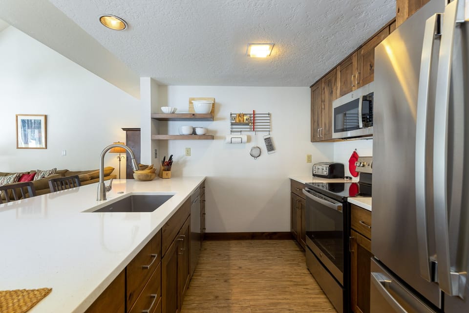 A modern kitchen with wooden cabinets, a white countertop, stainless steel appliances, and a double-door refrigerator. Open shelves hold dishes and a small toaster oven is on the counter.