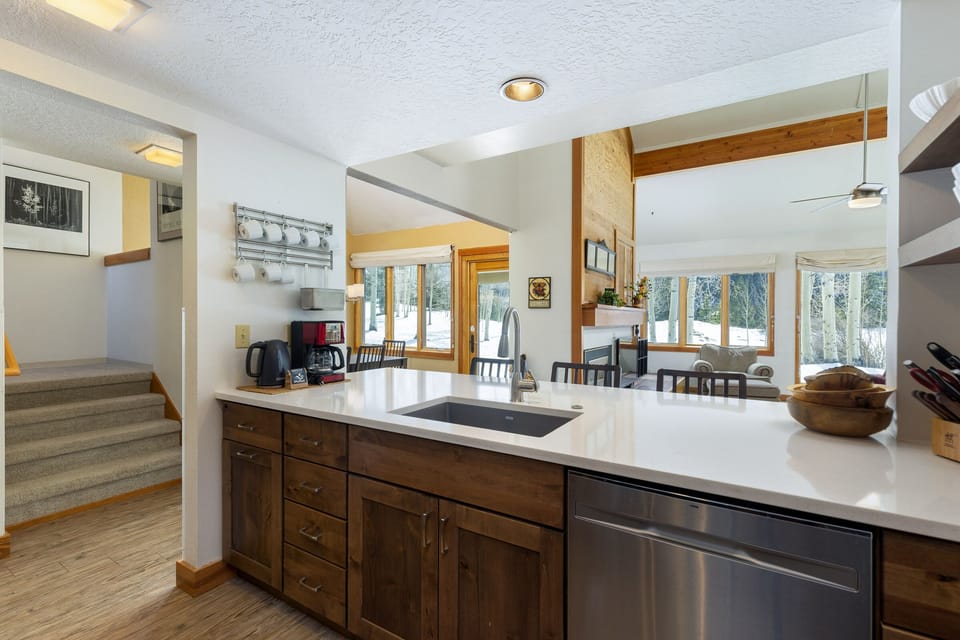 Modern kitchen with wood cabinets, white countertops, and stainless steel appliances. Open layout with a view of dining and living areas through large windows; staircase leading up visible on the left.