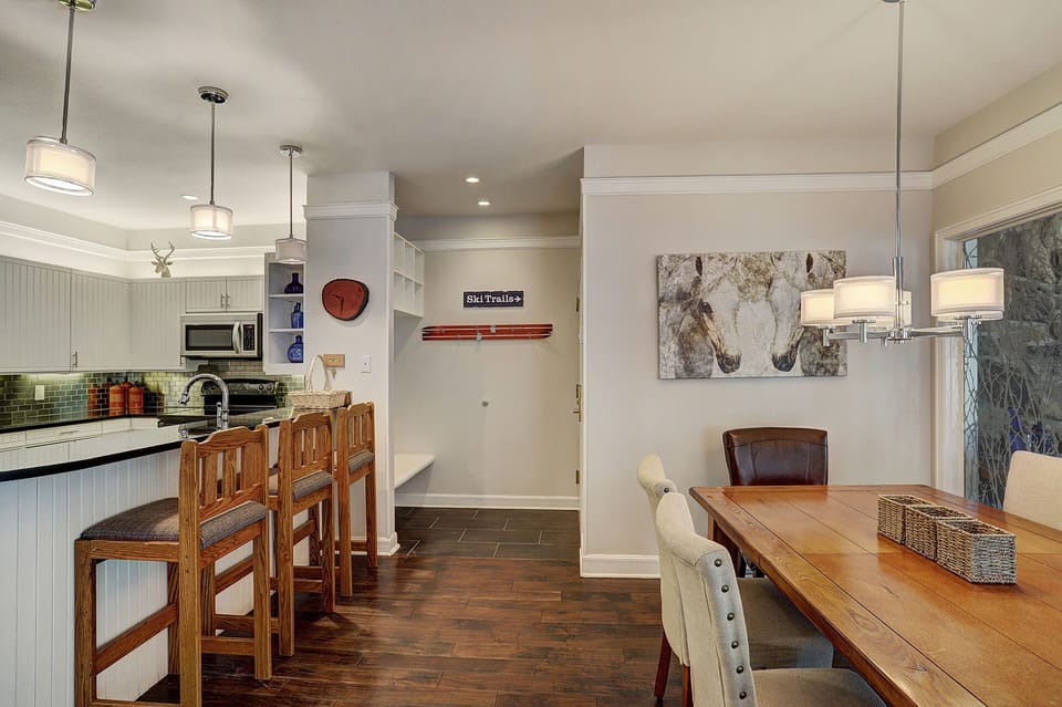 A modern kitchen and dining area with wooden bar stools, a dining table with chairs, pendant lighting, and artwork on the walls. The kitchen has white cabinets and stainless steel appliances.