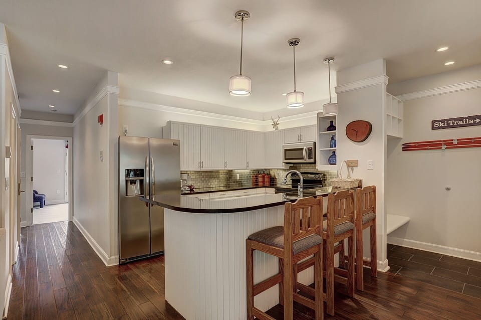 A modern kitchen with white cabinets, stainless steel appliances, a central island with wooden stools, and hardwood flooring. A hallway is visible to the left.