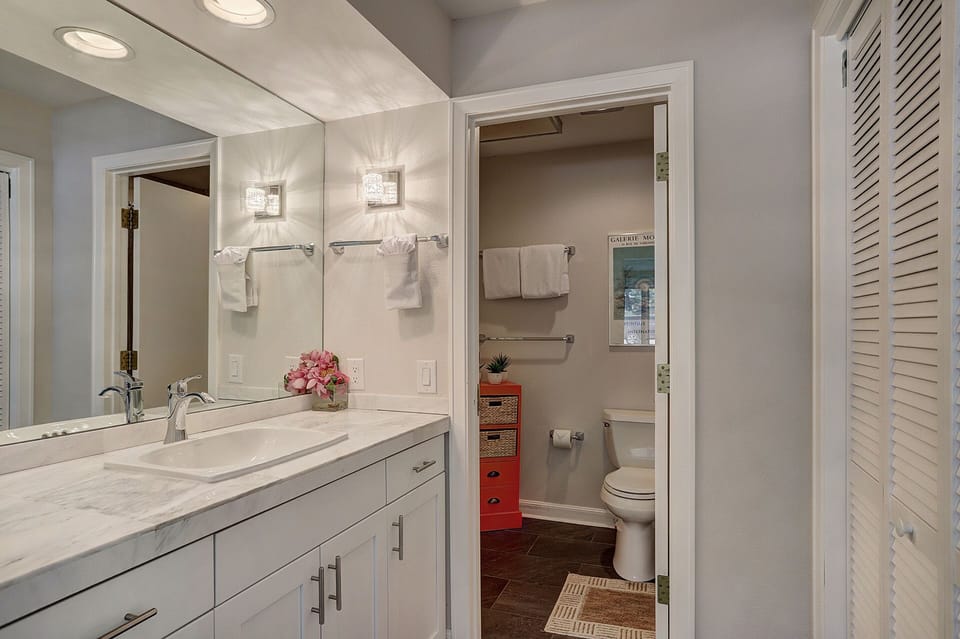 A modern bathroom with a sink and vanity mirror on the left, a toilet and shelving unit on the right, and a towel rack and wall art above the toilet.