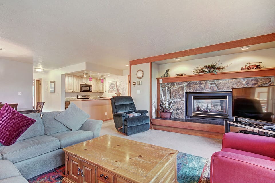 A cozy living room with a stone fireplace, gray sectional sofa, red armchair, wooden coffee table, and an open view of the kitchen area in the background.