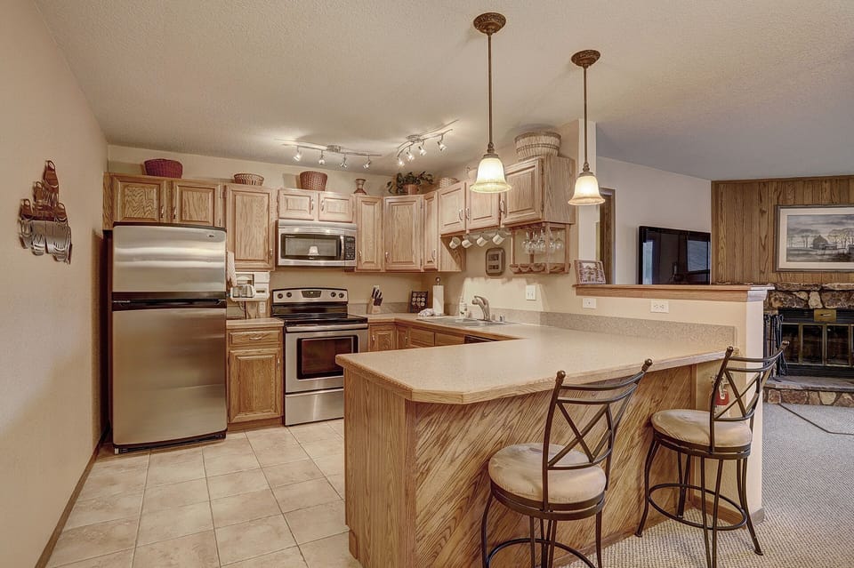 A kitchen with wooden cabinetry, stainless steel appliances, and tile flooring. The countertop extends to form a breakfast bar with two stools. Pendant lights hang above the counter.
