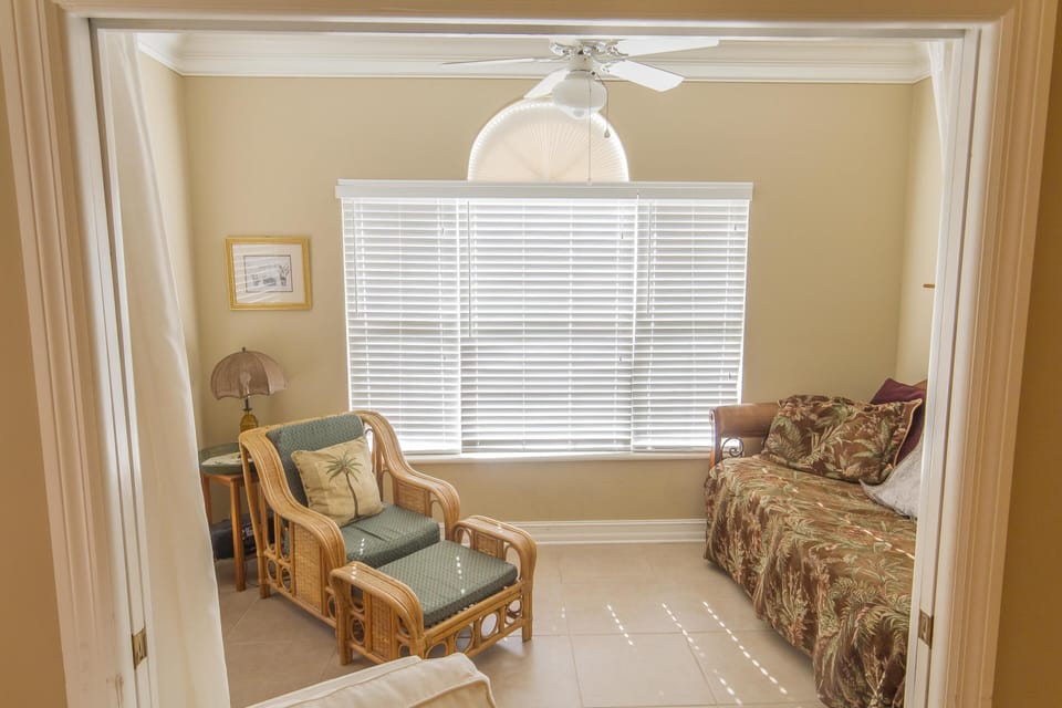 Sitting area with wicker armchair, tropical-patterned daybed, and white blinds filtering natural light.
