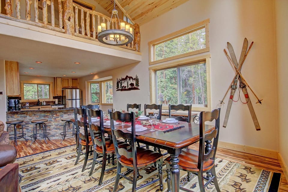A dining room features a wooden table with eight chairs, ski decor on the wall, large windows, and an adjoining open kitchen with bar stools and stone accents. Natural light fills the space.