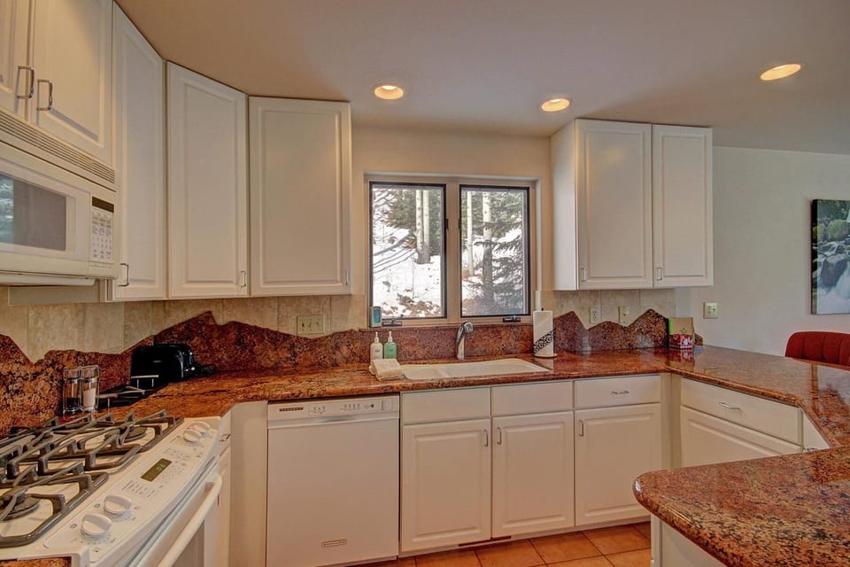 Modern kitchen with white cabinets, granite countertops, a dishwasher, stove, microwave, and sink. A window above the sink offers a view of trees and snow outside.
