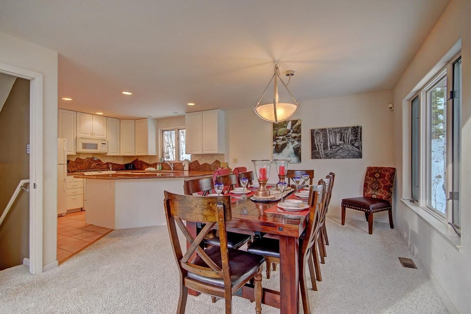 A dining area with a wooden table set for six beside a kitchen with white cabinets and modern appliances. A large window allows natural light, with forest-themed artwork on the walls.