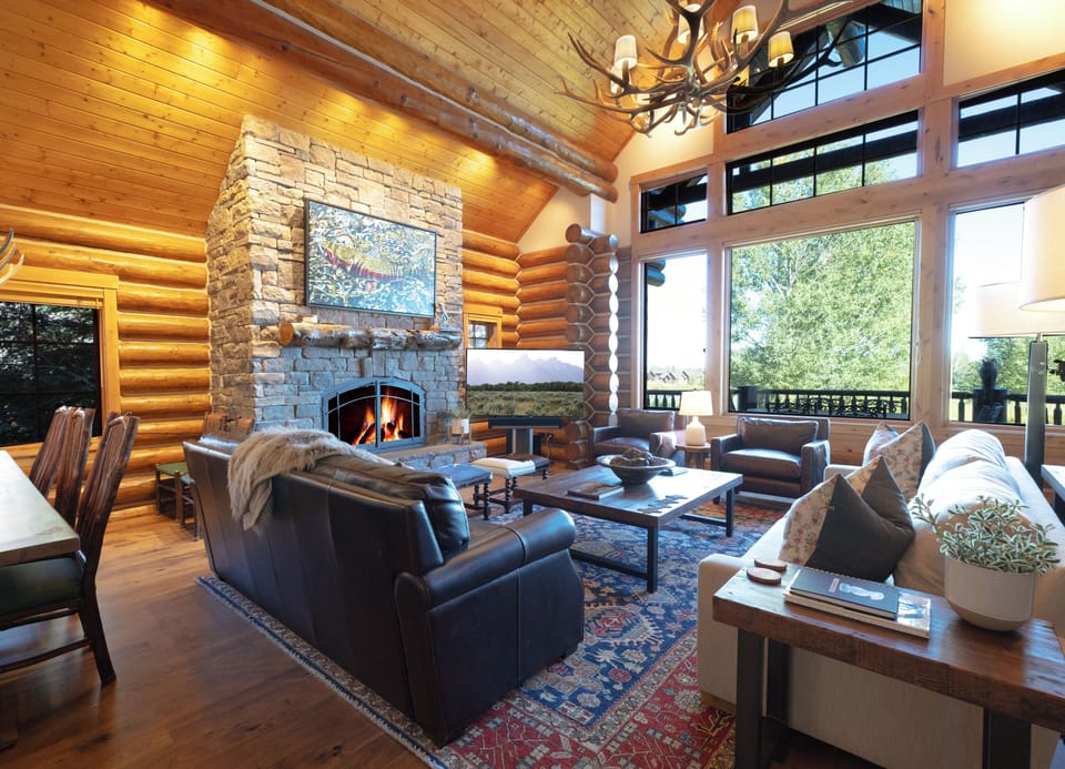 Curl up in your cozy mountain retreat where warm log walls and a crackling stone fireplace create the perfect ambiance for relaxing evenings with stunning forest views through soaring windows.