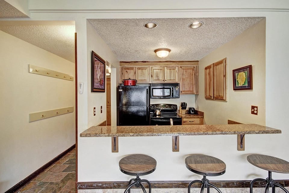 A compact kitchen with wooden cabinets, black appliances, and a granite countertop with three bar stools. Two framed artworks hang on the beige walls. The floor is tiled.
