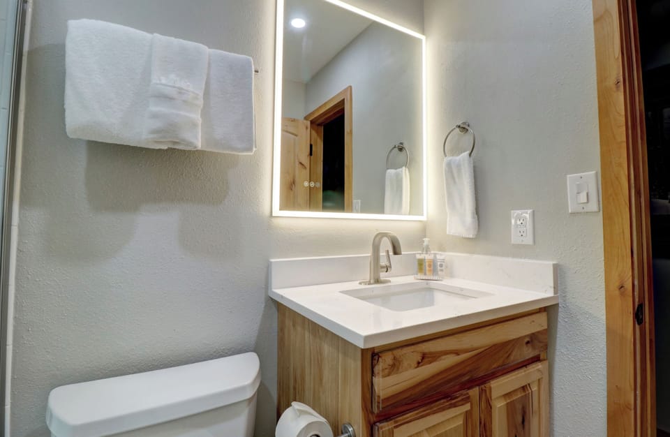 A small bathroom features a wooden vanity with a sink, an illuminated mirror, white towels on a ring holder and rack, and a toilet with a toilet paper roll holder.