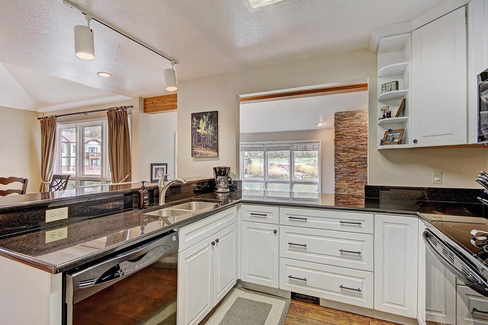 Modern kitchen with white cabinets, black countertops, stainless steel appliances, and a double sink. The space includes an adjacent dining area with large windows and beige curtains.