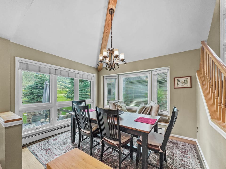 A dining room with a wooden table, black chairs, a patterned rug, and a chandelier. Large windows provide views of a green yard.