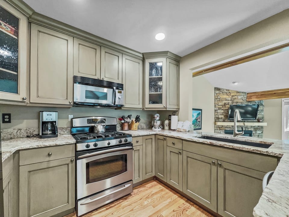 A modern kitchen with granite countertops, stainless steel appliances, beige cabinets, and a wood floor. An opening reveals a living area with a stone wall and TV.