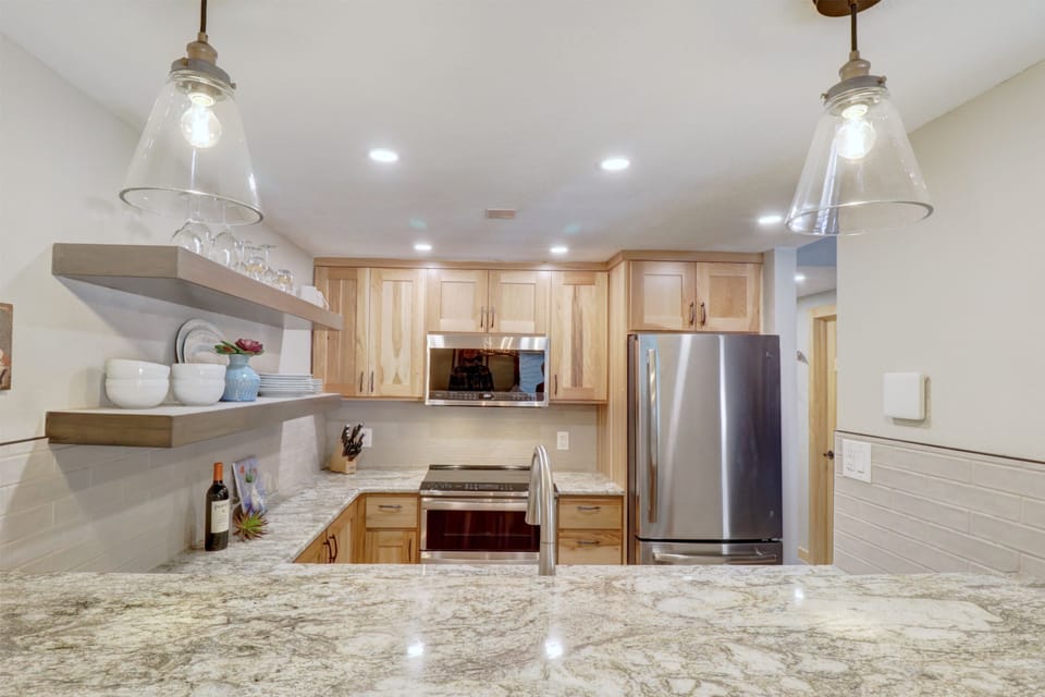 Modern kitchen with wooden cabinets, stainless steel appliances, open shelving, and granite countertops. Two pendant lights hang above the counter, and recessed lighting brightens the space.