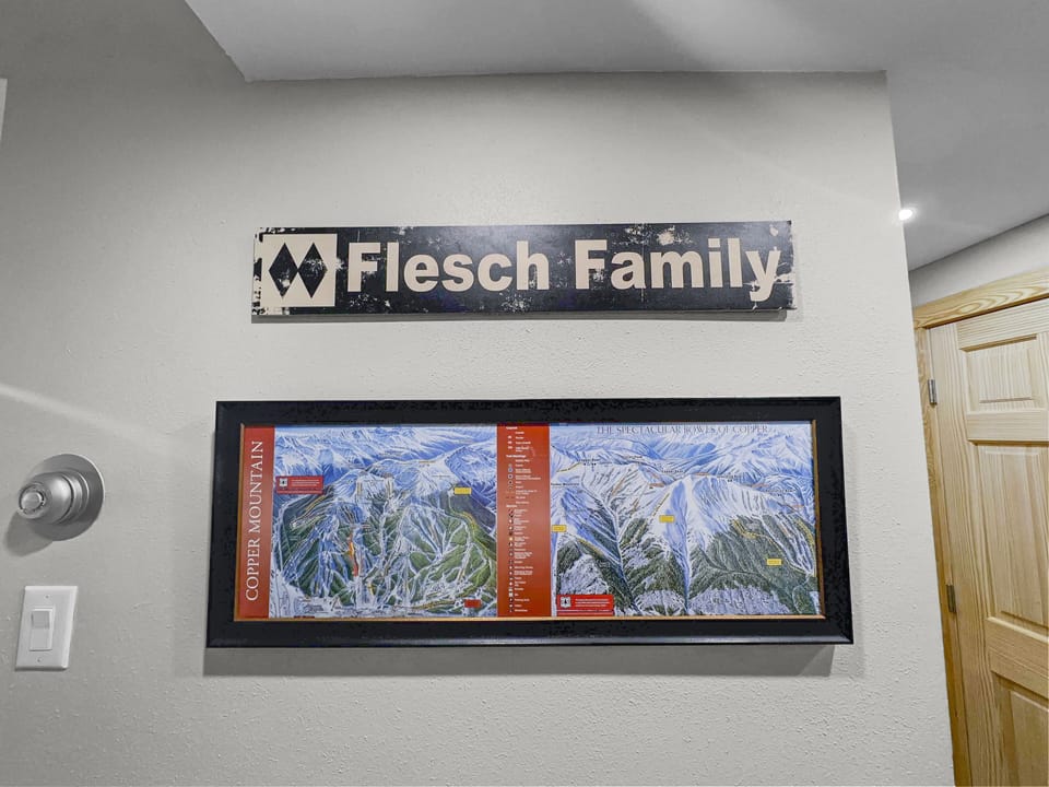 A sign reading "Flesch Family" is mounted above a framed map of Copper Mountain displayed on a beige wall. A wooden door and light switch are also visible.