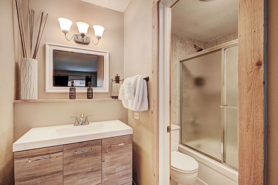 A bathroom with a sink, mirror, light fixture, and hand towels on one side, and a toilet with a glass-enclosed shower on the other side.