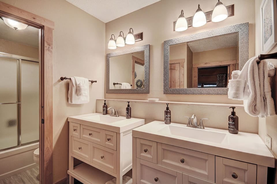 A modern bathroom with double sinks, large mirrors, and overhead lighting. It has a walk-in shower with a glass door and a neatly folded towel hanging by the counter.