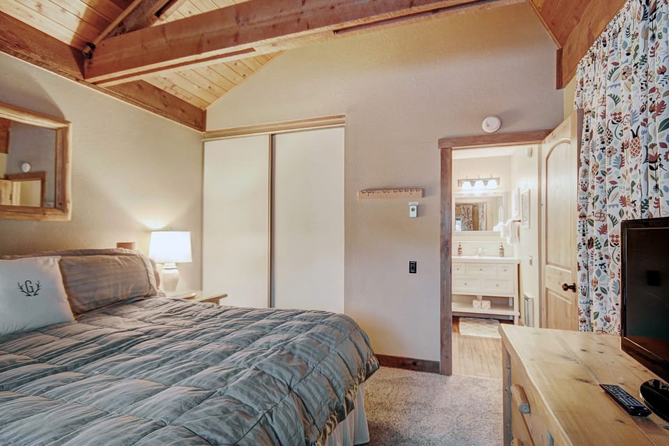 A cozy bedroom with a wooden ceiling has a bed with plaid bedding, a side lamp, a dresser with a TV, and an open doorway leading to a well-lit bathroom with a vanity and mirror.