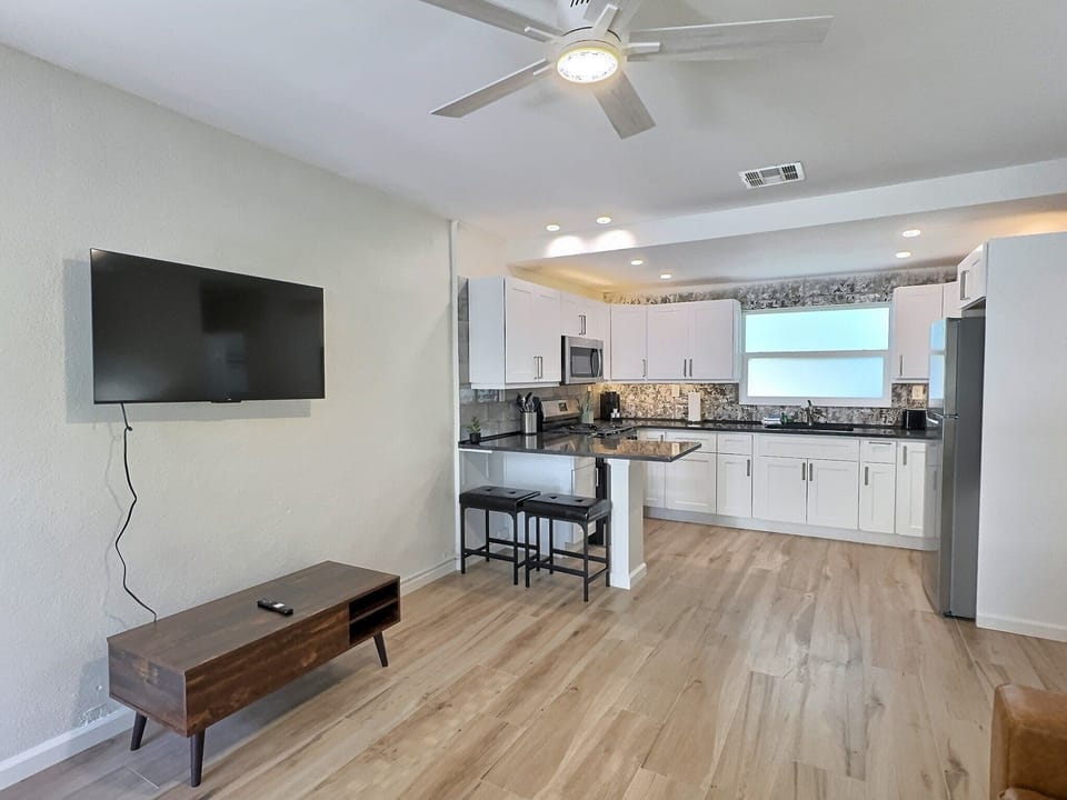 Living room with wall-mounted TV, sofa, coffee table, ceiling fan, and open kitchen
