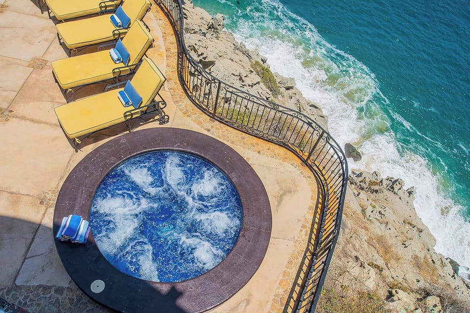 Aerial view of the property's dramatic oceanfront location, featuring a circular hot tub, yellow sun loungers on a curved terrace, and direct access to the rocky coastline and turquoise waters below.