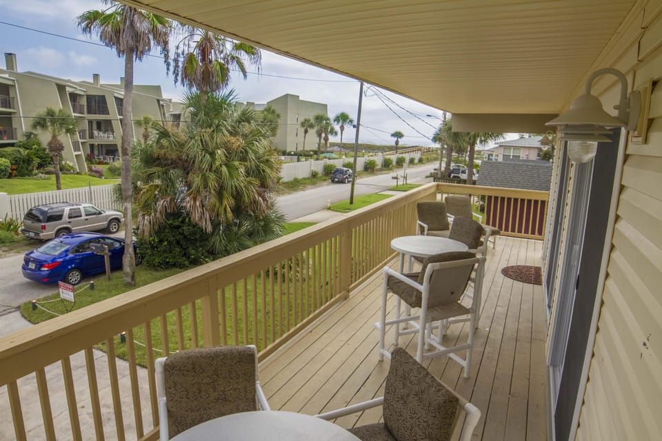 Upper balcony with cushioned chairs, round tables, and partial ocean view along a quiet street.