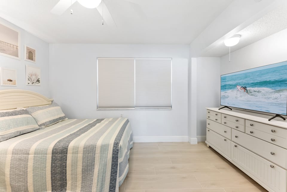 Bright bedroom featuring a comfortable bed, coastal-inspired decor, ceiling fan, and a dresser with flat-screen TV.