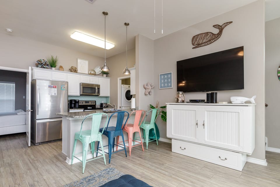 Bright open-concept space where your kitchen flows into the living area, featuring colorful bar stools and coastal charm for relaxed beachside dining.