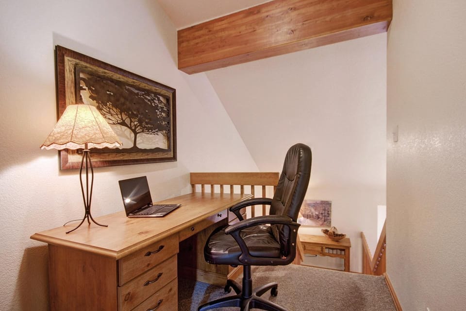 A home office with a wooden desk holding a laptop, a decorative lamp, and a black office chair. A framed painting hangs on the wall above the desk.
