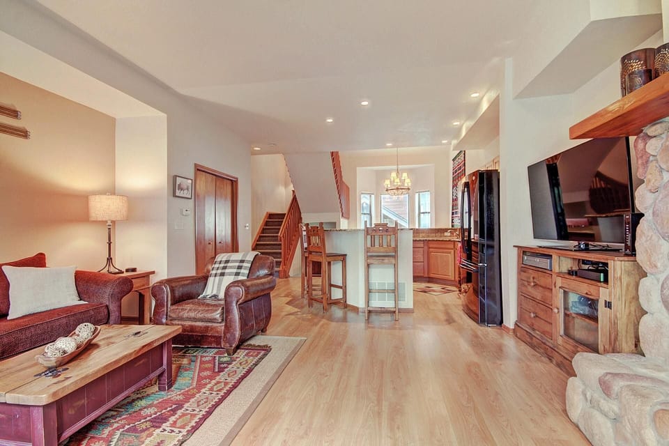 A cozy living room with wooden floors, a stone fireplace, comfortable seating, and a view of the adjacent dining area and kitchen in the background.