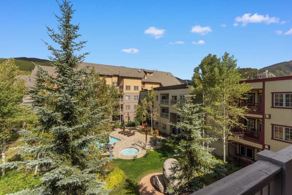 Mountain resort complex with pool and hot tub nestled among evergreen trees under clear blue skies.