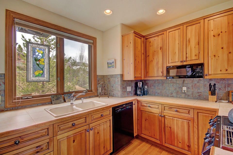 A kitchen with wooden cabinets, a large window with a stained glass decoration, a double sink, and a black dishwasher.