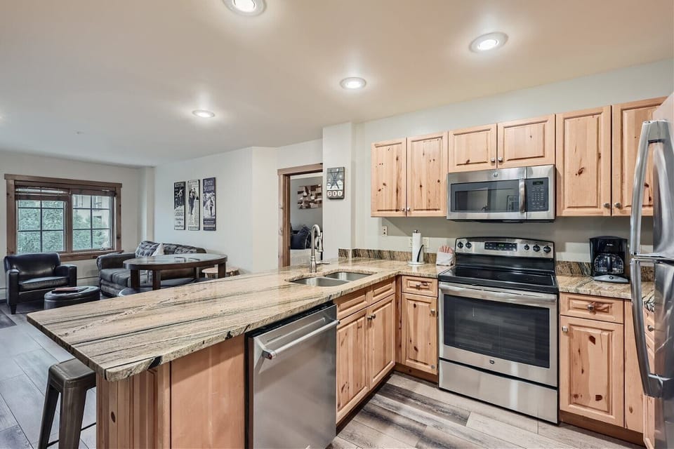 A modern kitchen with wooden cabinets, an island with a granite countertop, stainless steel appliances, and a view into the living area with a dining table and couch.