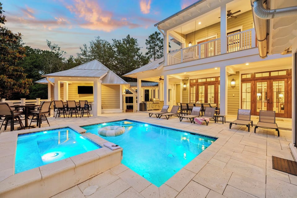 A beautiful vacation home featuring a private pool and spa surrounded by comfortable loungers, a covered outdoor dining pavilion with TV, and elegant two-story architecture nestled among towering pines.