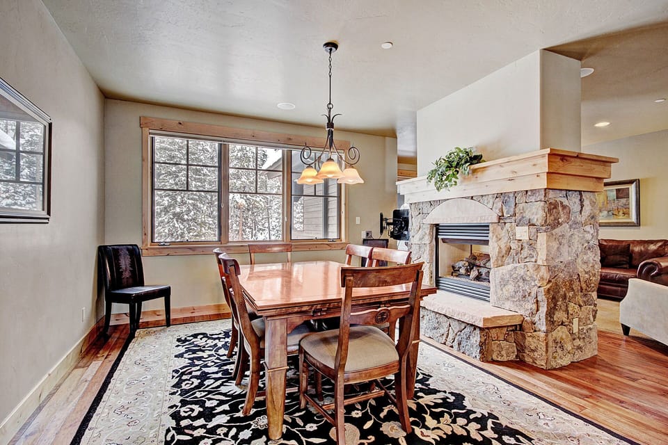 A dining room with a rectangular wooden table, four chairs, a light fixture above, a large window, and a stone fireplace with a mantel in a cozy interior space.