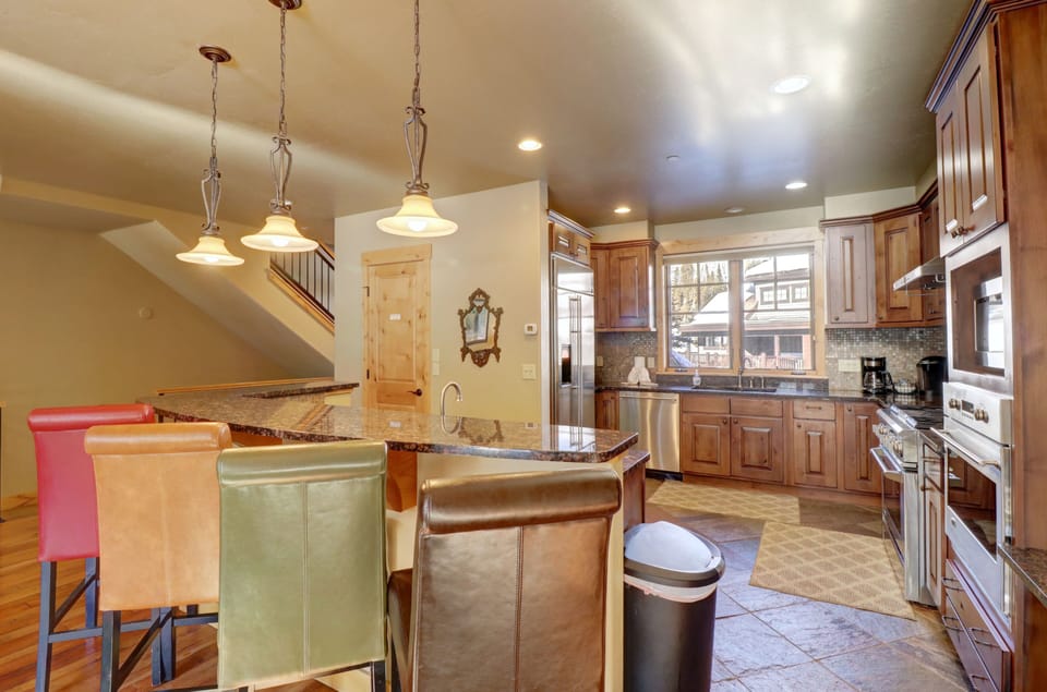 A spacious kitchen featuring wooden cabinets, stainless steel appliances, a granite island with pendant lights, and colorful bar stools.