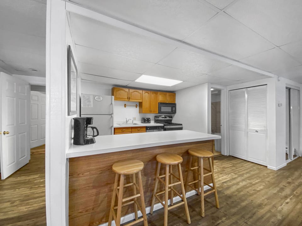 A small kitchen with wooden cabinets, countertop with three wooden stools, coffee maker, stove, microwave, and refrigerator. The floor is wooden, and there's a white door in the background.